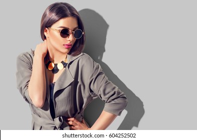 Beauty Fashion brunette model girl wearing stylish coat and sunglasses. Sexy woman portrait with perfect makeup, trendy accessories and fashion wear. Beauty trends. Isolated on grey. Fashion blogger
