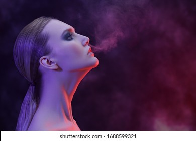 Beauty and fashion concept. Art portrait of a gorgeous woman with dark make-up smoking in red light on a black background. 