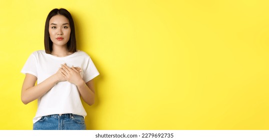 Beauty and fashion concept. Beautiful asian woman holding hands on heart and looking thoughtful at camera, keeping memories in soul, standing over yellow background. - Shutterstock ID 2279692735