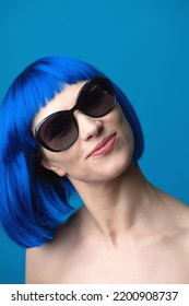 Beauty, Fashion Concept. Portrait Of Beautiful And Sexy Woman With Blue Wig And Sunglasses Looking To Camera With Smile. Sun Tan Soft Skin, Pink Lipstick And White Teeth. Model With Naked Shoulders