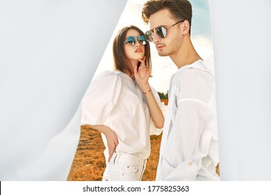 Beauty, Fashion. Couple Of Fashionable Young People Posing Outdoor In White Clothes And Sunglasses Among White Fabrics Fluttering In The Wind. Summer Style. Sunlight.