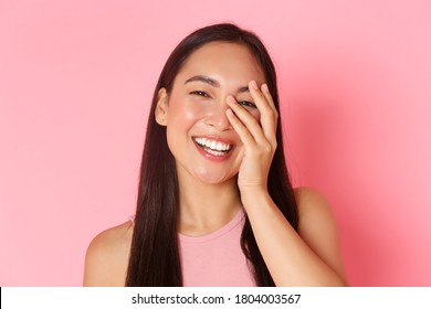 Beauty, fashion and lifestyle concept. Close-up of carefree beautiful asian woman touching face and laughing happily with closed eyes, standing over pink background joyful, skincare product promo