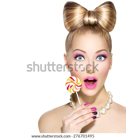 Beauty fashion model girl Eating colourful lollipop. Lollypop. Surprised Young funny woman with bow hairstyle, pink nail art and makeup isolated on white background