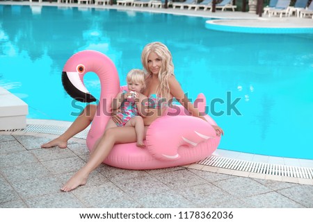 Beauty fashion mother with daughter family look. Beautiful blond woman with having fun with little pretty gil wears in swim wear posing on Inflatable Flamingo Pool Float by swimming pool. 
