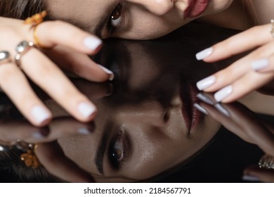 Beauty, Fashion, Style And Make-up Concept. Studio Portrait Of Beautiful Woman With Fancy Make-up Lying On Black Glass Surface And Looking To Camera. Model Face And Hands Reflection On Dark Mirror