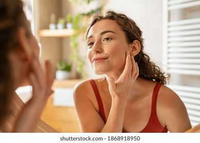 Beauty girl looking at mirror while touching her face and checking pimple, wrinkles and bags under the eyes, during morning beauty routine. Happy smiling beautiful young woman applying moisturizer.  - Shutterstock ID 1868918950
