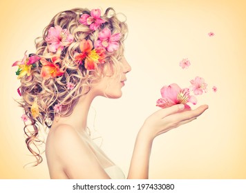 Beauty girl takes beautiful flowers in her hands. Blowing flower. Hairstyle with flowers.  Fantasy girl portrait in pastel colors. Summer fairy portrait. Long permed hair.