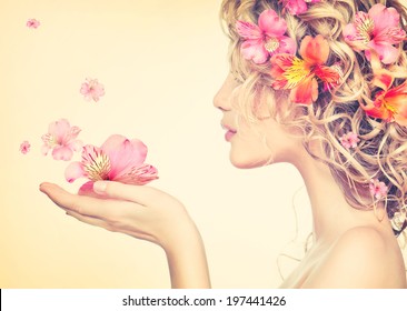 Beauty girl takes beautiful flowers in her hands. Blowing flower. Hairstyle with flowers.  Fantasy girl portrait in pastel colors. Summer fairy portrait. Long permed hair.
