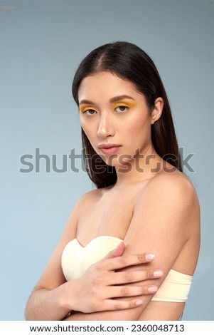beauty and grace, asian model with bold makeup and bare shoulders posing on blue backdrop