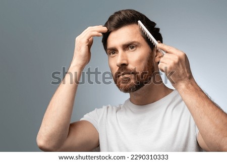 Beauty, grooming and hairstyle concept. Handsome caucasian bearded middle aged man brushing hair with comb, posing over gray studio background