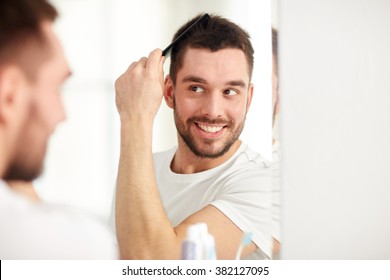 Beauty, Grooming And People Concept - Smiling Young Man Looking To Mirror And Brushing Hair With Comb At Home Bathroom