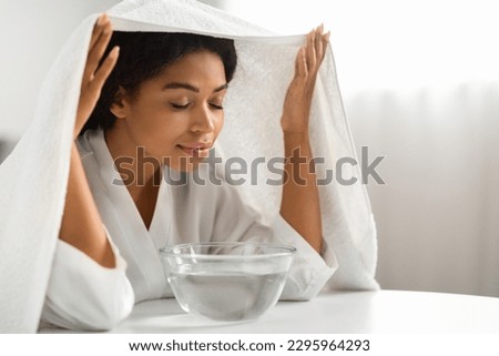Beauty Hacks. Young black woman inhalling steam of hot water at home, beautiful african american female sitting at table and breathing warm air coming from bowl, covered with bath towel, closeup