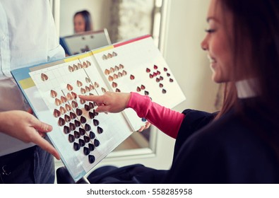 Beauty, Hair Dyeing And People Concept - Happy Young Woman With Hairdresser Choosing Hair Color From Palette Samples At Salon