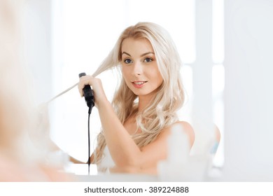 Beauty, Hairstyle, Morning And People Concept - Smiling Young Woman With Styling Iron Straightening Her Hair And Looking To Mirror At Home Bathroom