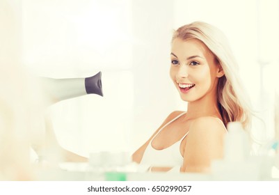 Beauty, Hairstyle, Morning And People Concept - Smiling Young Woman With Fan Blow Drying Her Hair Looking To Mirror At Home Bathroom