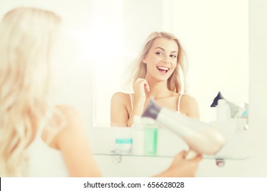 Beauty, Hairstyle, Morning And People Concept - Smiling Young Woman With Fan Blow Drying Her Hair Looking To Mirror At Home Bathroom