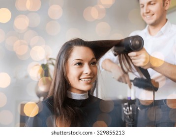 beauty, hairstyle and people concept - happy young woman and hairdresser with fan making hot styling at hair salon over holidays lights