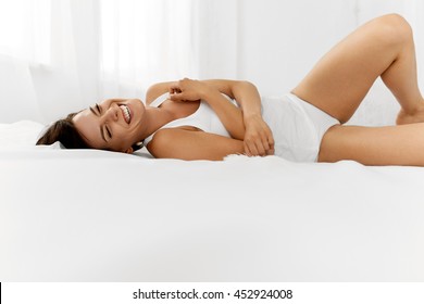 Beauty And Health. Beautiful Smiling Woman With Fresh Soft Skin And Natural Makeup In Underwear Having Fun Lying On White Bed. Healthy Happy Female Model Relaxing Indoors. Body And Skin Care Concept