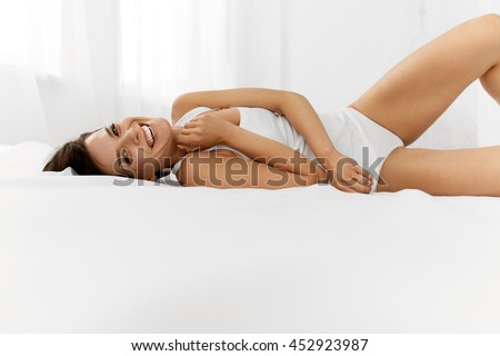 Beauty And Health. Beautiful Smiling Woman With Fresh Soft Skin And Natural Makeup In Underwear Having Fun Lying On White Bed. Healthy Happy Female Model Relaxing Indoors. Body And Skin Care Concept