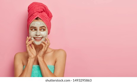 Beauty and health care concept. Gentle positive woman applies facial mask, pampers skin after taking shower, touches cheeks gently, isolated over pink background. Face care and cosmetology concept