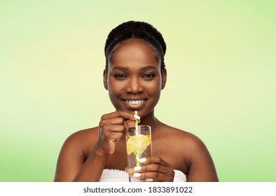 Beauty, Healthy Eating And People Concept - Portrait Of Smiling African American Woman With Bare Shoulders Drinking Lemon Water From Glass With Paper Straw Over Lime Green Natural Background