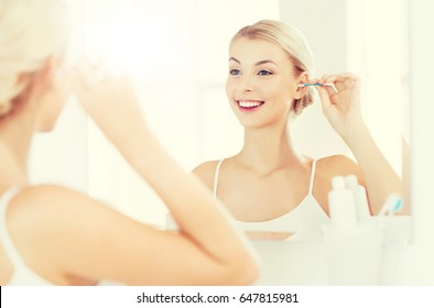 美しさ、衛生、人々のコンセプト – 笑顔の若い女性が綿棒で耳を掃除し、自宅のトイレで鏡を見る