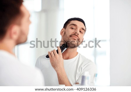 beauty, hygiene, shaving, grooming and people concept - young man looking to mirror and shaving beard with trimmer or electric shaver at home bathroom