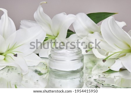 Beauty jar cream cosmetic with lily flowers lie on table, jar cream product on wet flower mirror moistening. Natural flower jar cream cosmetics for hand skin care. Smooth beauty skin