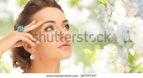 beauty, jewelry, people and\
accessories concept - close up of woman face with cocktail ring on\
hand and earrings over summer garden and cherry blossom\
background