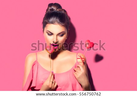 Beauty joyful Young fashion model Girl eating Valentine Heart shaped cookies on sticks in her hands. Love Concept. Beautiful seductive young woman. Valentines Day gift. On pink background