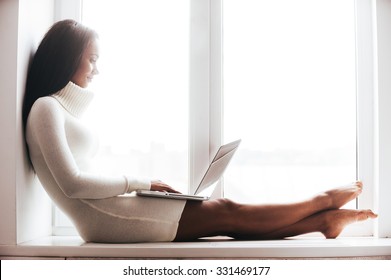 Beauty With Laptop. Attractive Young African Woman In Warm Sweater Working On Laptop While Sitting On The Window Sill