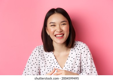 Beauty and lifestyle concept. Close-up of happy asian woman laughing and having fun, standing over pink background