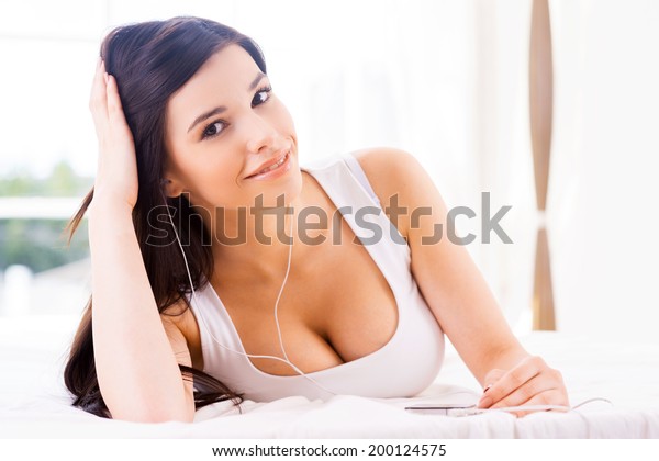 Beauty listening to the music. Beautiful young\
smiling woman listening to the MP3 player and looking at camera\
while lying in bed