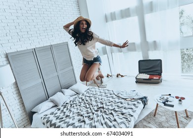 Beauty in the mid-air. Full length of playful young woman in sun hat smiling while jumping on the bed