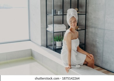 Beauty model with healthy perfect skin applies cream for body care, poses wrapped in towel after taking shower, poses at bathtube, looks thoughtfully away, feels refreshed and relaxed. Spa procedures - Powered by Shutterstock