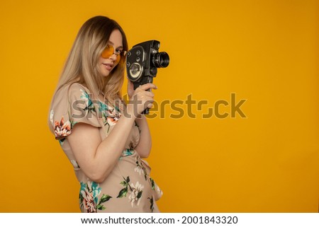 Beauty with movie camera. Cheerful young woman holding movie camera and posing against yellow background