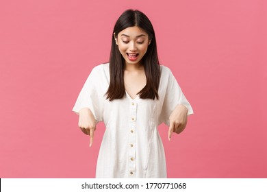 Beauty, people emotions and summer leisure and vacation concept. Surprised and excited kawaii asian girl in white dress, pointing and looking down with amused happy face, pink background