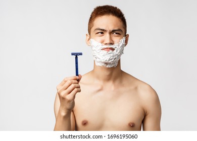 Beauty, People And Hygiene Concept. Portrait Of Reluctant And Displeased Asian Man With Naked Torso, Looking Away, Dont Want To Shave But Need, Holding Razor And Apply Facial Cream With Uneasy Face