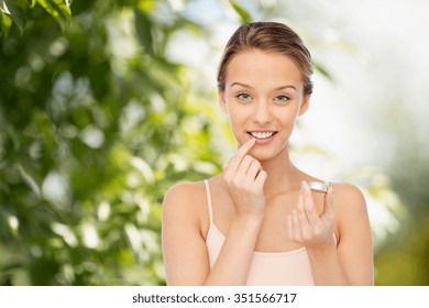 Beauty, People And Lip Care Concept - Smiling Young Woman Applying Lip Balm To Her Lips Over Green Natural Background