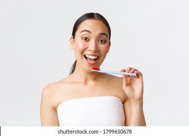 Beauty, Personal Care, And Hygiene Concept. Enthusiastic Happy Asian Woman In Bath Towel Getting Ready, Brushing White Teeth, Holding Toothbrush And Smiling Broadly, White Background