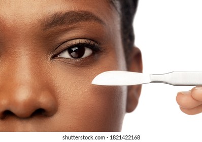 Beauty, Plastic Surgery And People Concept - Close Up Of Face Of Beautiful Young African American Woman And Hand With Scalpel Knife Over White Background