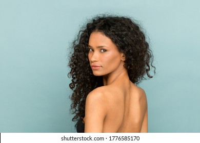 Beauty Portrait Of A Afro American Woman With Gorgeous Long Curly Hair Turning To Look Back Over Her Shoulder At The Camera Against A Blue Studio Background