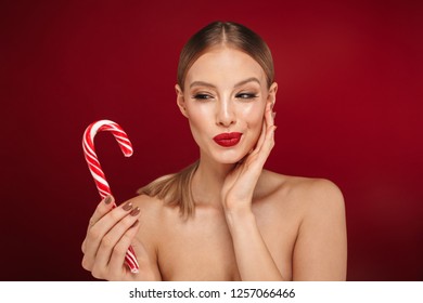 Beauty portrait of an attractive blonde haired topless woman standing isolated over red background, eating Christmas candy