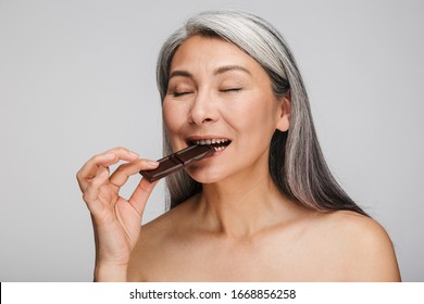 Beauty Portrait Of An Attractive Mature Topless Woman With Long Gray Hair Standing Isolated Over Gray Background, Eyes Closed, Eating Dark Chocolate