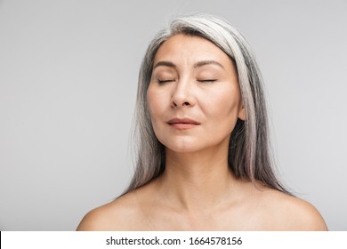 Beauty portrait of an attractive sensual mature topless woman with long gray hair standing isolated over gray background, eyes closed