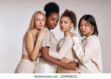 beauty portrait of diverse females, caucasian, asian and african american ladies with different skintone posing at camera, hugging each other, dressed casually in beige tone clothes. isolated