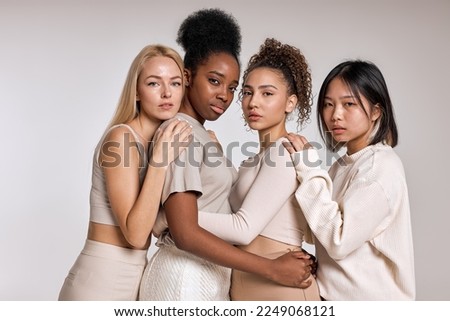 beauty portrait of diverse females, caucasian, asian and african american ladies with different skintone posing at camera, hugging each other, dressed casually in beige tone clothes. isolated