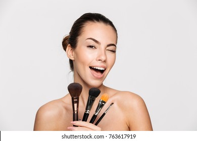 Beauty portrait of an excited beautiful half naked woman holding set of make-up brushes and winking isolated over white background
