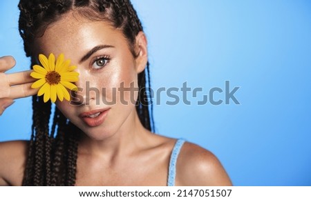 Beauty portrait of fashionable young woman with summer dreads hairstyle, tanned healthy facial skin, clear face, showing flower, standing over blue background