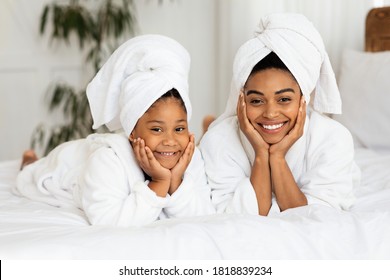 Beauty Portrait Of Happy Black Mom And Daughter In Bathrobes And Towels On Head Lying On Bed At Home, Posing After Bath, Having Spa Day, Cute Little Girl And Her Mother Having Fun Together, Free Space